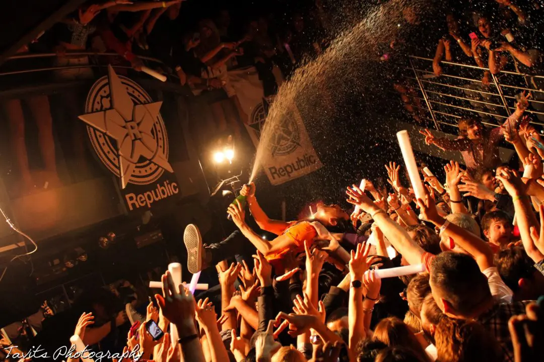 A performer crowd surfs at a packed concert, spraying liquid from a bottle over cheering fans. Hands reach up, and bright stage lights illuminate the energetic atmosphere—unlike any typical corporate event or seminar. "Republic" banners hang on the walls.