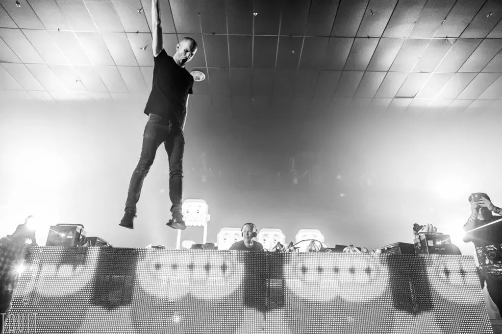 A performer hangs by one arm from the ceiling above a DJ booth at a vibrant corporate event, with bright lights and a smoky atmosphere behind him. Several people are visible behind the booth, and the crowd is partially visible in the foreground.