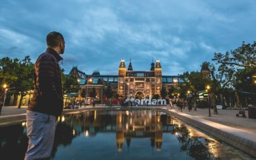 A man stands near a reflective pool at dusk, facing the illuminated Rijksmuseum in Amsterdam, where a corporate event is taking place beside the "I amsterdam" sign.