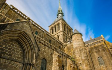 A low-angle view of Mont Saint-Michel Abbey in France, its tall spire and arched windows soaring—an inspiring backdrop for a seminar or corporate event beneath a bright blue sky with wispy clouds.