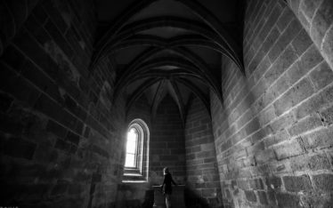 A person stands alone under vaulted stone ceilings in a dark, empty seminar room, gazing out of a single arched window where daylight streams in, illuminating their silhouette.