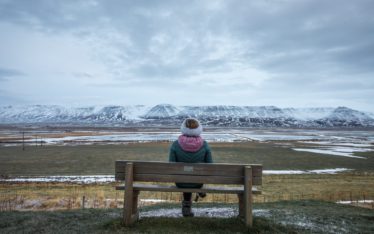 A person in a winter coat and hat sits alone on a wooden bench, reflecting quietly after a corporate event, facing a vast snowy landscape with distant mountains under a cloudy sky.