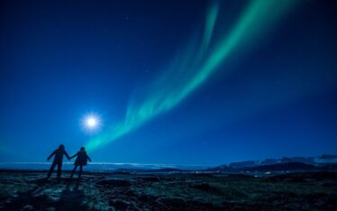 Two people holding hands stand on a rocky landscape at night, gazing at the moon and vibrant green Northern Lights, as if sharing a quiet seminar beneath the clear, starry sky.