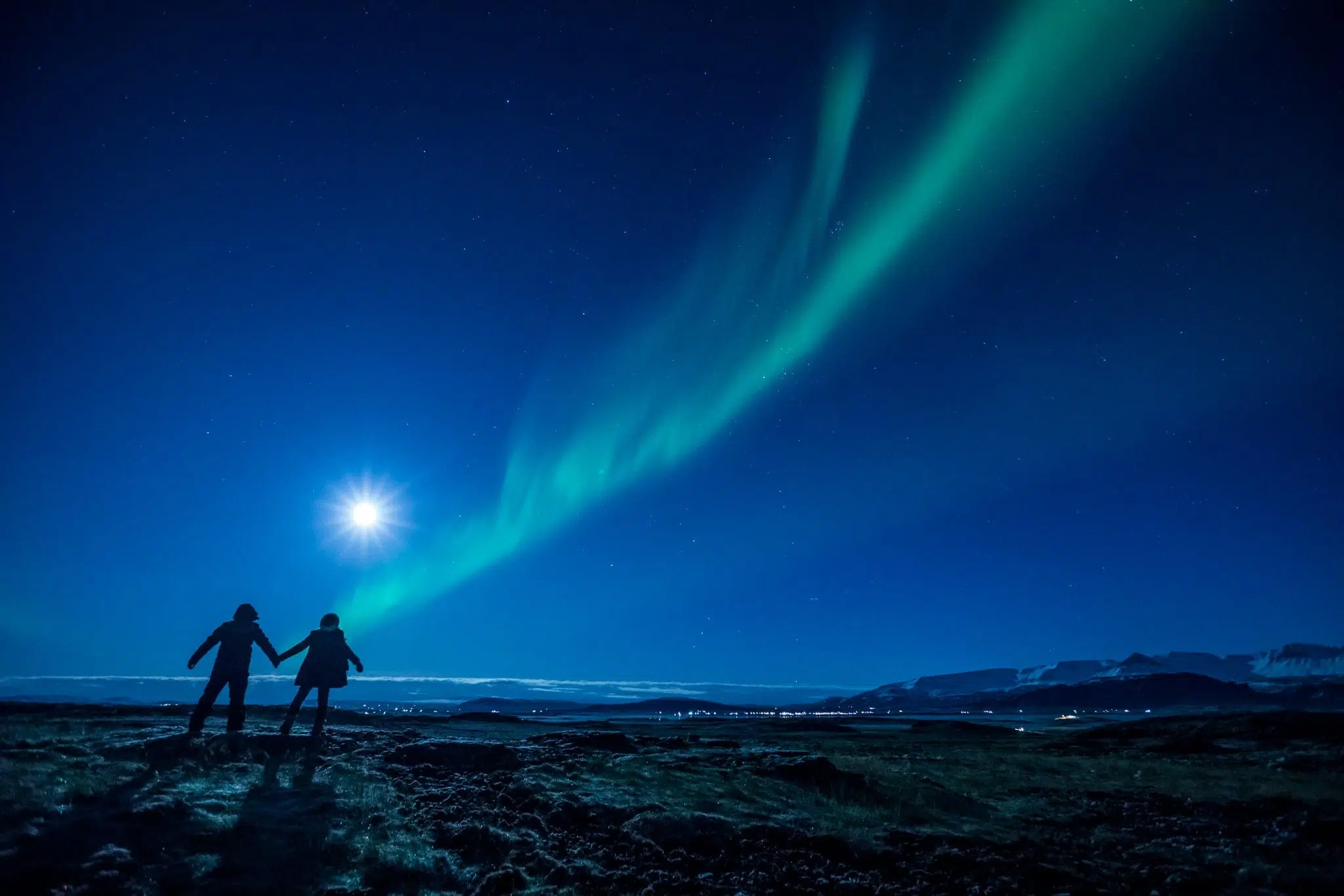 Two people holding hands stand on a rocky landscape at night, gazing at the moon and vibrant green Northern Lights, as if sharing a quiet seminar beneath the clear, starry sky.