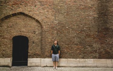 A man in a dark shirt and shorts stands against a large, cracked brick wall with a black arched door on the left—perhaps pausing near the site of a former corporate event—along a quiet cobblestone street.