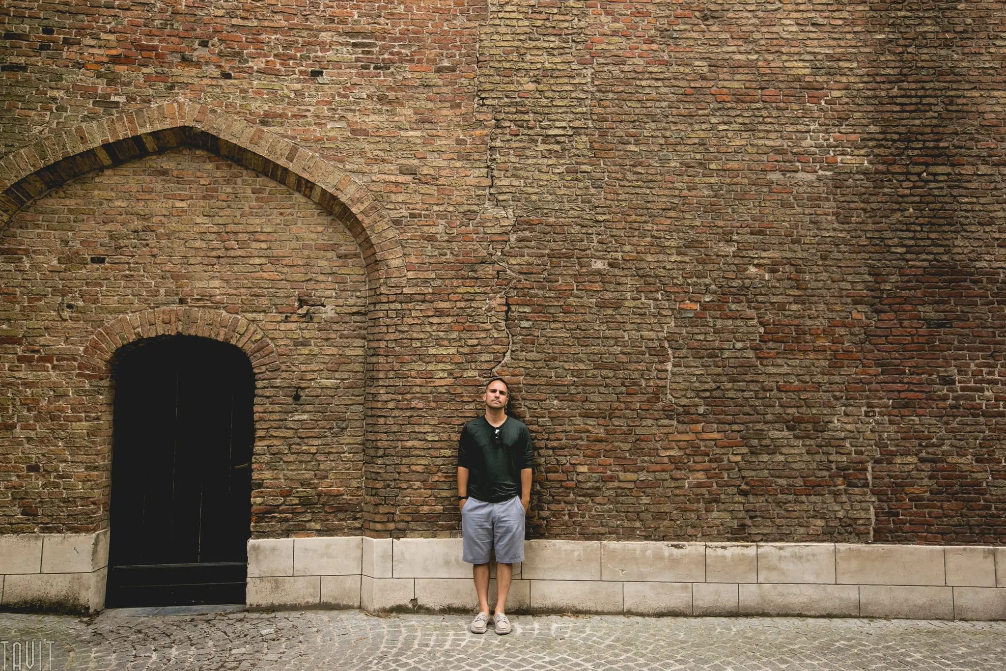 A man in a dark shirt and shorts stands against a large, cracked brick wall with a black arched door on the left—perhaps pausing near the site of a former corporate event—along a quiet cobblestone street.