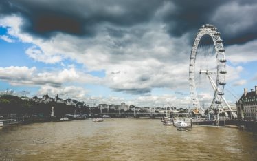 The image shows the London Eye ferris wheel beside the River Thames under a dramatic sky with dark clouds. Several boats are on the river and historic buildings line the riverbank, making it an inspiring backdrop for a corporate event or seminar.