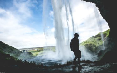 Silhouette of a person standing in a cave behind a large waterfall, with mist, green hills, and blue sky visible through the water—an inspiring backdrop perfect for a memorable corporate event or seminar.