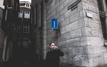 A man in casual clothes stands on a street corner beside a tall stone building, drinking from a can near a dead-end sign. The narrow street and surrounding architecture create an urban atmosphere, far removed from any corporate event or seminar setting.