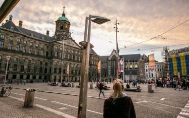 A person stands in a large cobblestone square at sunset, facing a historic building with a green dome. People gather for a seminar, with banners and streetlights illuminating the scene and creating an inviting atmosphere.