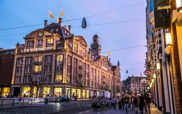 Historic De Bijenkorf department store in Amsterdam at dusk, its windows aglow. People walk and cycle by, some heading to a nearby seminar or corporate event amid the lively street lined with shops and illuminated signs under a blue-purple sky.
