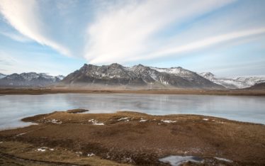 A wide, shallow lake with patches of snow and brown grass in the foreground, backed by rugged, snow-dusted mountains under a pale blue sky with streaks of white clouds—an inspiring setting perfect for a corporate event.