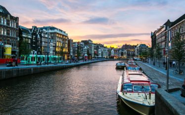 A canal in a European city at sunset, lined with traditional buildings, boats docked along the water, and people walking on the sidewalk—perfect for an evening stroll after a corporate event or seminar. The sky is colorful with clouds and warm sunlight.