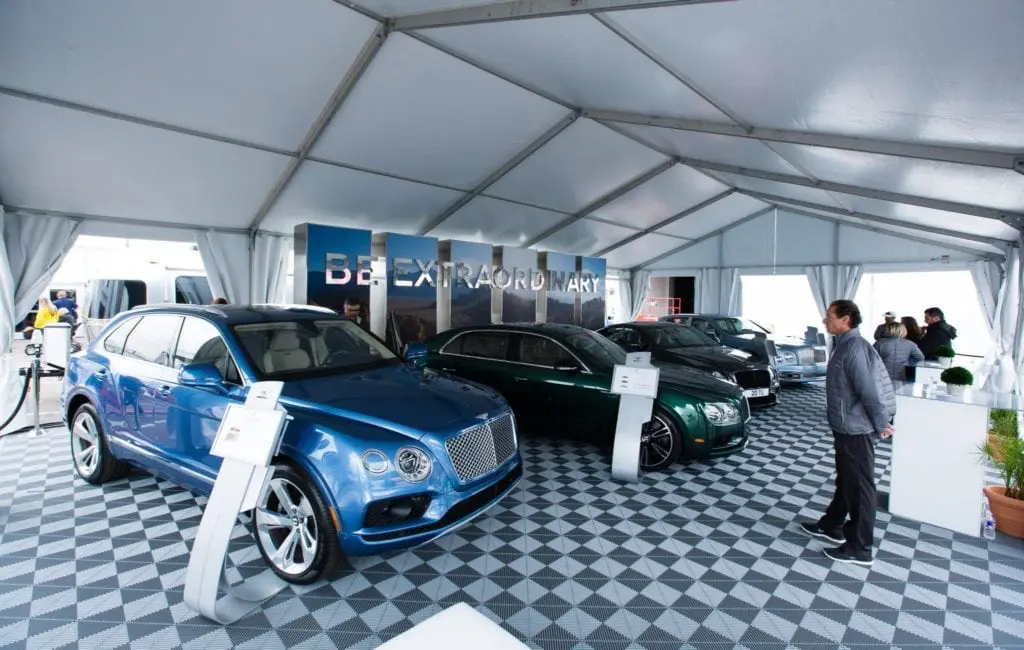 A man stands inside a large white tent at a corporate event displaying luxury Bentley cars, including a blue SUV and green sedan, on a checkered floor with informational signs in front of each car. Several people observe the vehicles.