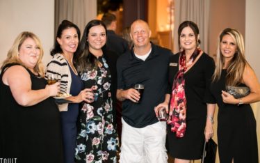 Six adults, four women in dresses and one man in a black shirt and white shorts, stand together smiling at a corporate event, holding drinks and posing for a group photo at an indoor seminar.