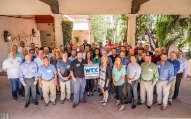 A large group of people, both men and women, stand outdoors in a courtyard, smiling at the camera during a corporate event. The group is holding a sign that reads "WTX" with "World Trade Exchange" written beneath it.