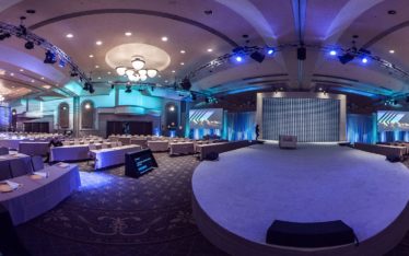 A spacious, elegant conference hall set for a seminar or corporate event, with rows of tables and chairs facing a large stage, enhanced by blue lighting and chandeliers—perfect for formal presentations.
