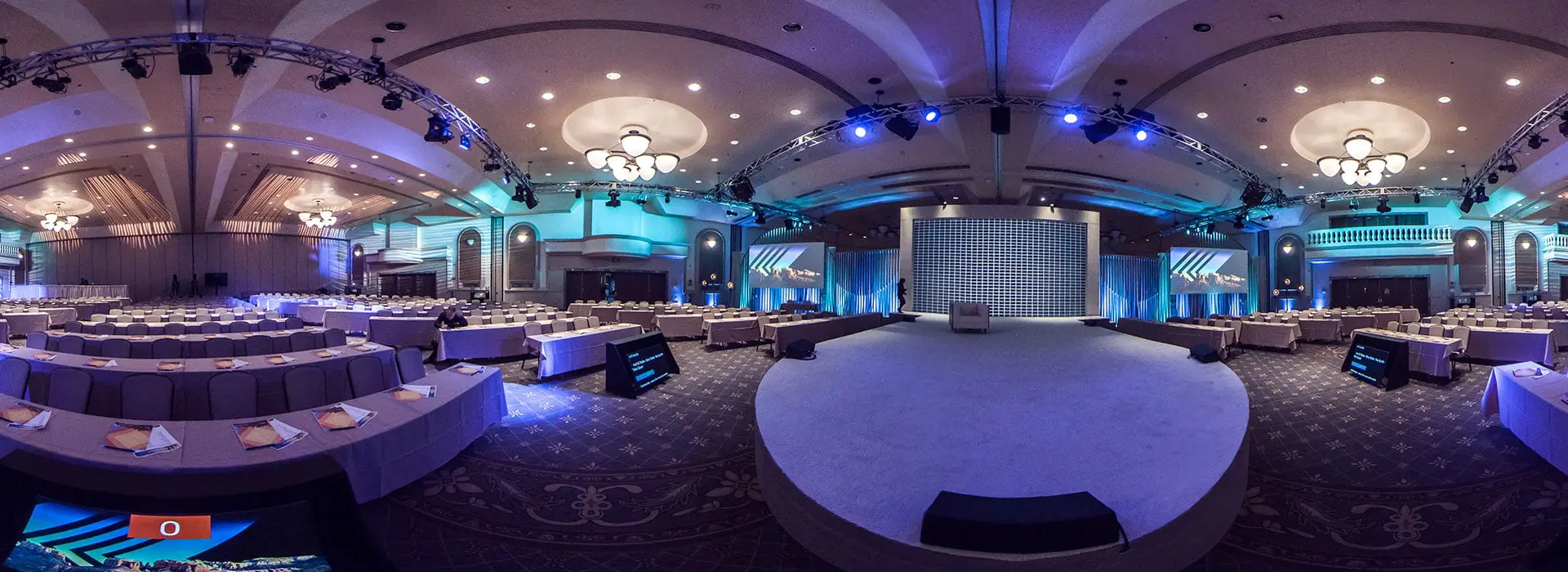 A spacious, elegant conference hall set for a seminar or corporate event, with rows of tables and chairs facing a large stage, enhanced by blue lighting and chandeliers—perfect for formal presentations.