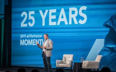 A man in a suit stands on stage speaking at a corporate event in front of a blue screen displaying "25 YEARS" and "2019 Manhattan MOMENTUM." Two white chairs and a small table with water are also on stage.