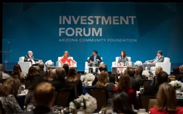 A panel of five speakers sit on stage at the Arizona Community Foundation Investment Forum seminar, facing an audience. The stage backdrop displays the event name in large letters, highlighting this prominent corporate event.