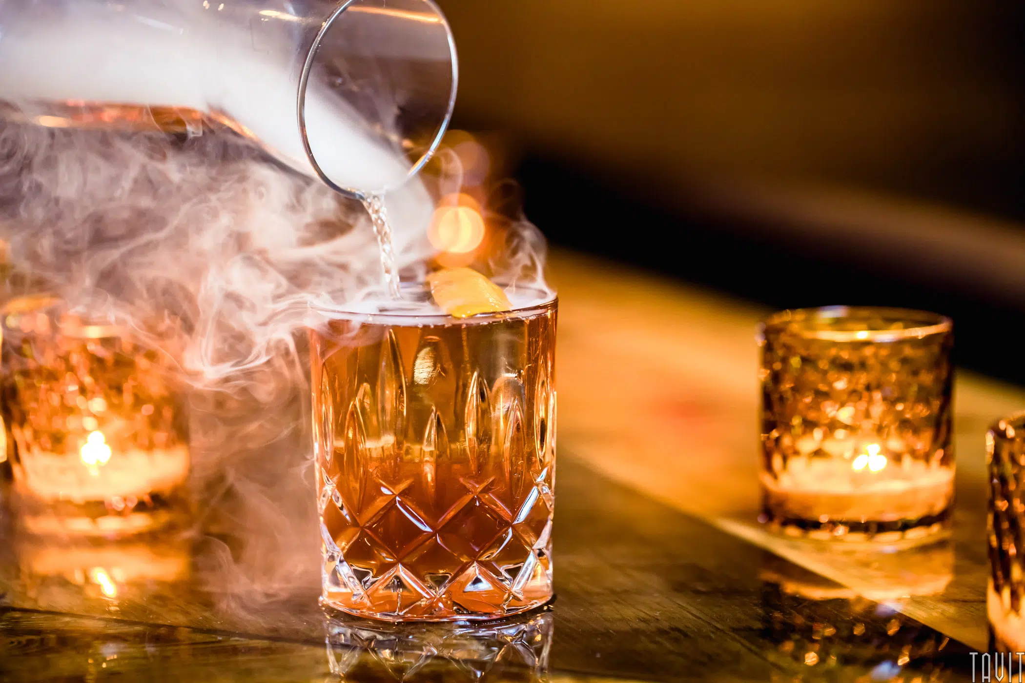 A glass featuring a decorative pattern showcases an amber-hued cocktail garnished with an orange twist. Smoke swirls dramatically as a smoky liquid is expertly poured from a small pitcher, captured in stunning product photography against the candlelit ambiance.