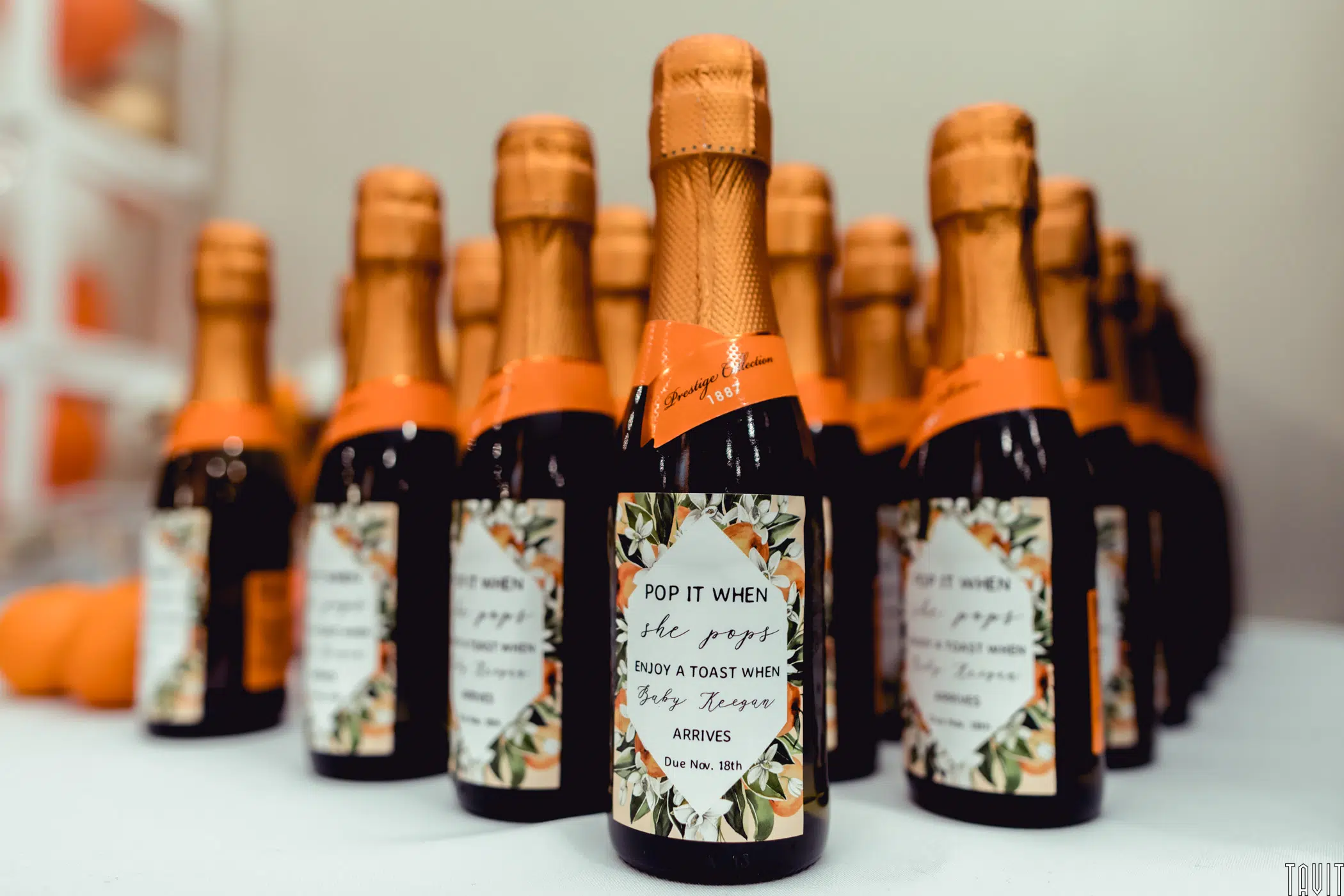 Rows of small champagne bottles with orange foil tops and decorative labels, perfect for a seminar or corporate event, read “Pop it when she pops. Enjoy a toast when baby Russo arrives. Due Nov. 18th.” The bottles are arranged on a table.