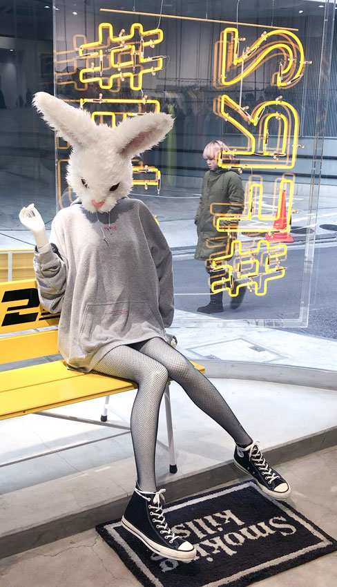 Woman in bunny mask sitting on bench in front of neon sign