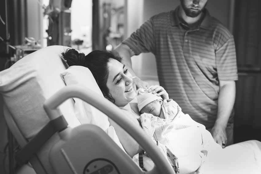 Mother in hospital bed holding new baby father standing black and white