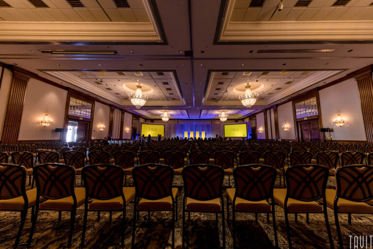 A large, elegant conference room set up for a corporate event or seminar, with rows of empty chairs facing a stage lit by yellow screens and blue lighting; chandeliers and ornate decor complete the sophisticated expo atmosphere.
