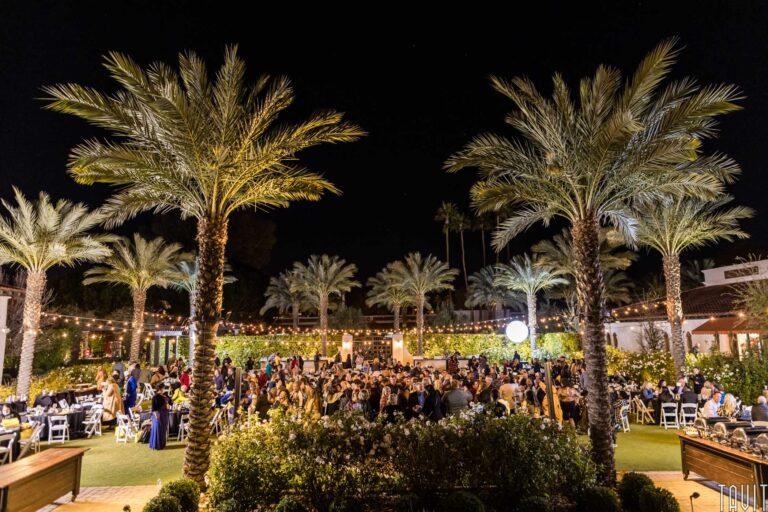 An outdoor corporate event with many people seated at tables under string lights, surrounded by tall palm trees and lush greenery, creating a festive and elegant atmosphere.