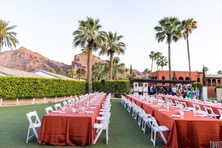 Outdoor corporate event setup with long tables covered in red tablecloths and white chairs, surrounded by palm trees and mountains. People are gathered near a building in the background under a clear sky, perfect for a seminar.