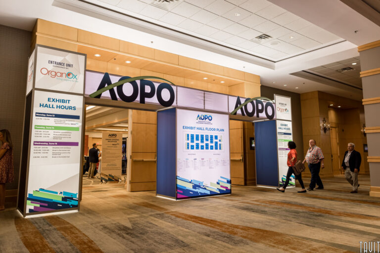 Convention center entrance with AOPO signage welcomes attendees to the seminar, with the exhibit hall floor plan and exhibit hours posted. Several people walk in and out of the brightly lit hallway, reflecting a modern corporate event atmosphere.