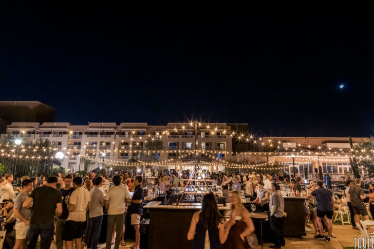 A lively crowd gathers outdoors at night under string lights for a corporate event, with people talking and milling around food and drink stands. Modern buildings are illuminated in the background against a dark sky.