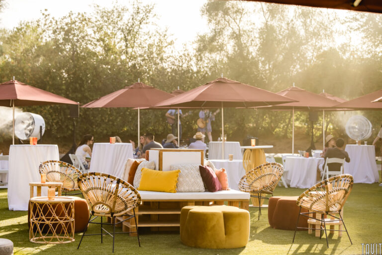 Outdoor corporate event with people sitting at round tables covered in white cloths, shaded by large red umbrellas. In the foreground, wicker chairs and a white couch with colorful pillows create a relaxed seminar atmosphere on green grass.