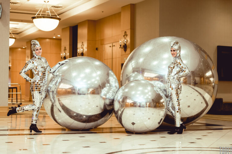 Two people in shiny, mirrored bodysuits and headpieces pose playfully beside three large reflective silver spheres at a sleek corporate event in a modern, well-lit indoor space with marble floors and chandeliers.