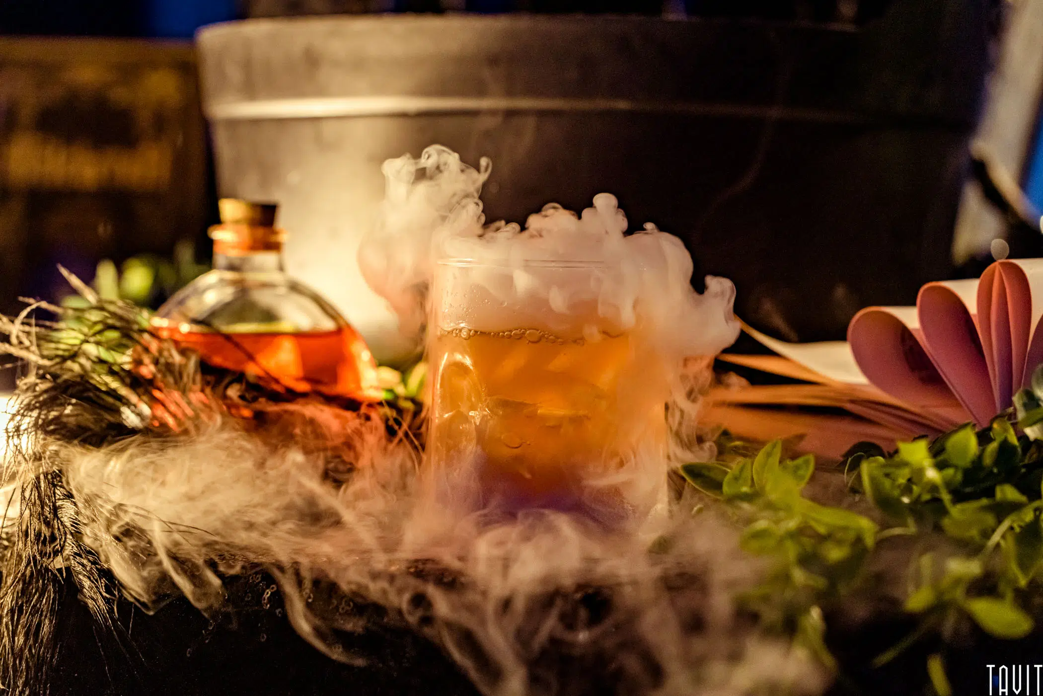 A glass filled with a smoky cocktail sits on a table surrounded by green leaves and a round bottle with amber liquid, making it the perfect centerpiece for a seminar or corporate event, with vapor dramatically swirling around the drink.