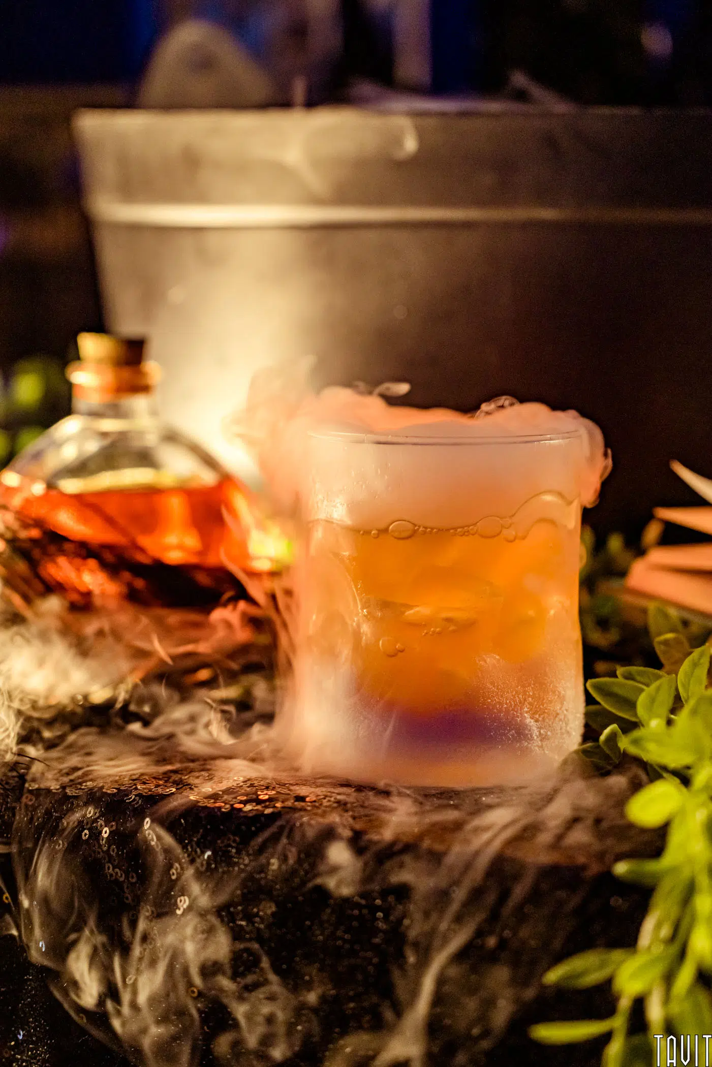 A frosty glass of orange drink emits vapor, surrounded by mist, with a decorative bottle of liquor and green leaves nearby—perfect for adding elegance to any corporate event or seminar set against a dark, moody background.