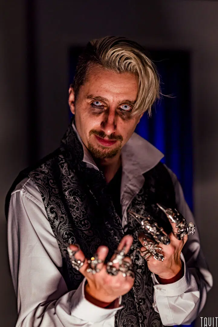 A man with slicked-back blond hair, pale makeup, dark eye circles, and a goatee stares intensely. Dressed for a corporate event, he wears a patterned vest, white shirt, and multiple large silver rings with claw-like accessories on his fingers.