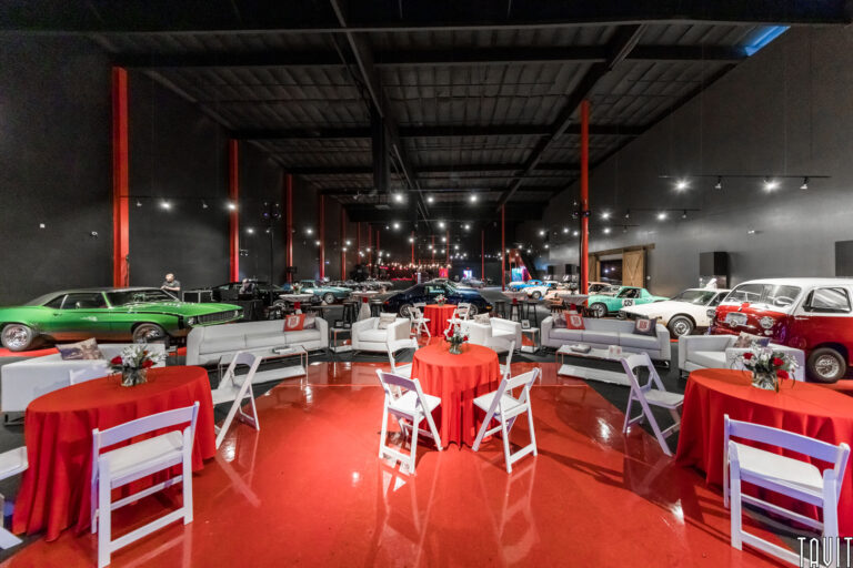 A spacious venue perfect for a corporate event or seminar, featuring classic cars on display, red floors, and white chairs and tables adorned with red tablecloths and floral centerpieces.