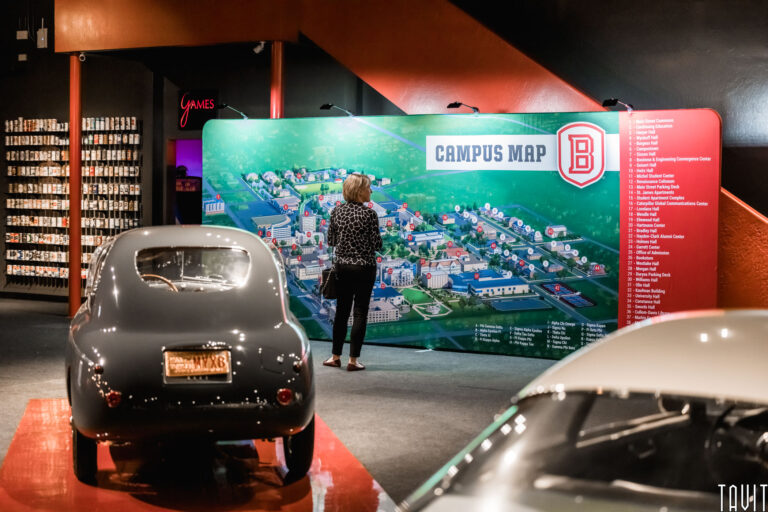 A person stands in front of a large, colorful campus map display indoors at a seminar, with vintage cars visible in the foreground and shelves labeled “Games” in the background.