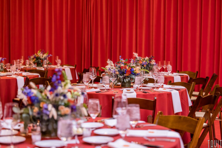 Round tables with red tablecloths and white napkins are set for a formal corporate event, decorated with colorful flower centerpieces, surrounded by wooden chairs, and placed in front of a bright red curtain.