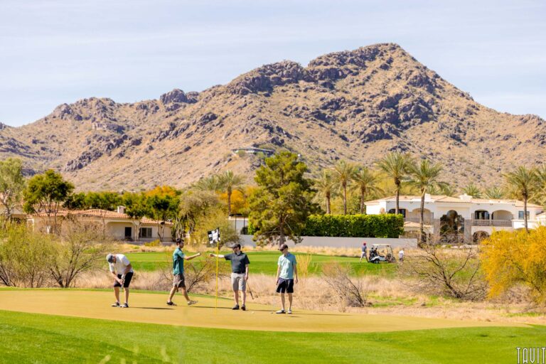 Four people are playing golf on a green surrounded by desert trees and shrubs. Mountains rise under a blue sky. In the distance, two white houses and golf carts suggest a scenic spot for a seminar or corporate event.