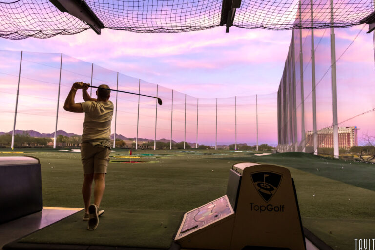 A person is swinging a golf club at a TopGolf driving range during sunset, with a purple and pink sky, mountains in the distance, and nets surrounding the field at a lively corporate event.