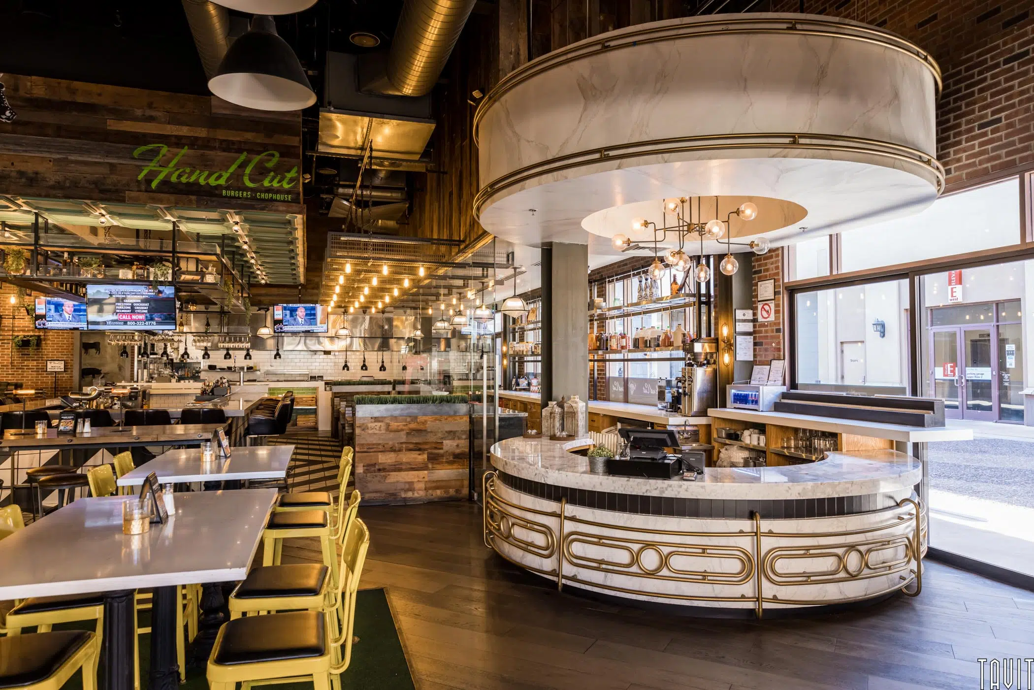 Modern restaurant interior with a round marble bar, shelves of bottles, wooden accents, yellow chairs, and large windows—perfect for hosting a seminar or corporate event in a stylish space with both natural and contemporary lighting.