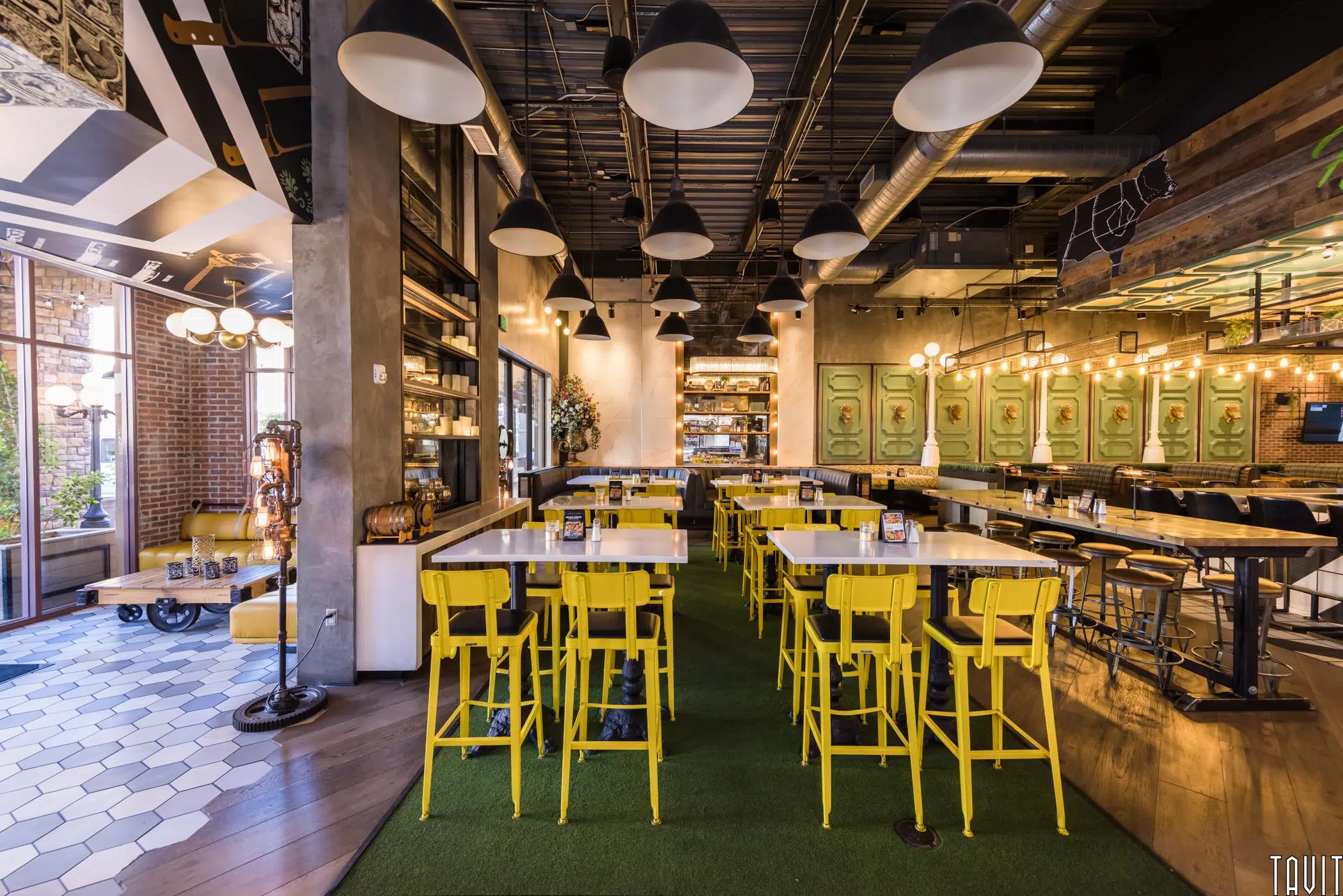 A modern restaurant interior with yellow chairs around white tables, green carpeting, and eclectic decor—ideal for hosting a seminar or corporate event thanks to its spacious layout, exposed ceilings, and large windows allowing natural light.