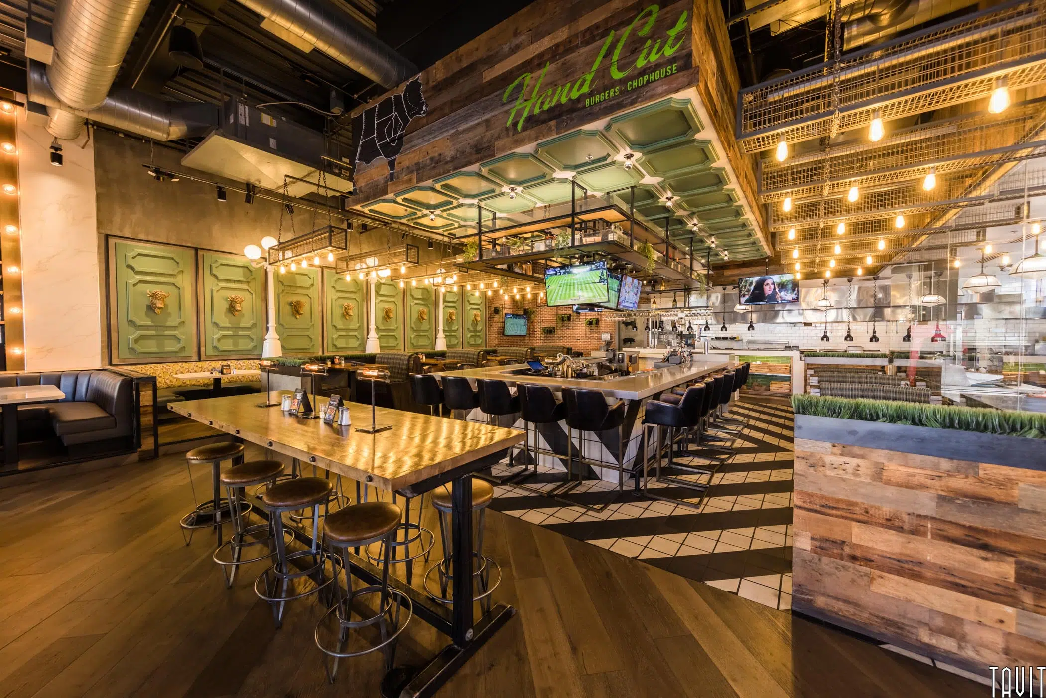 A stylish, modern restaurant interior features a central bar with high stools, wood and metal tables, black booths, hanging lights, and green decorative panels—perfect for hosting a corporate event in a warm and inviting atmosphere.