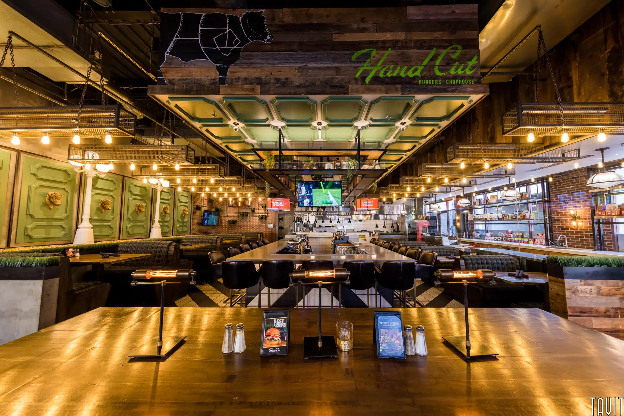 Modern restaurant interior with wood and metal décor, hanging lights, booth seating, a long bar, TV screens, and “Hand Cut” signage with a cow diagram above—an inviting atmosphere ideal for a corporate event or seminar.