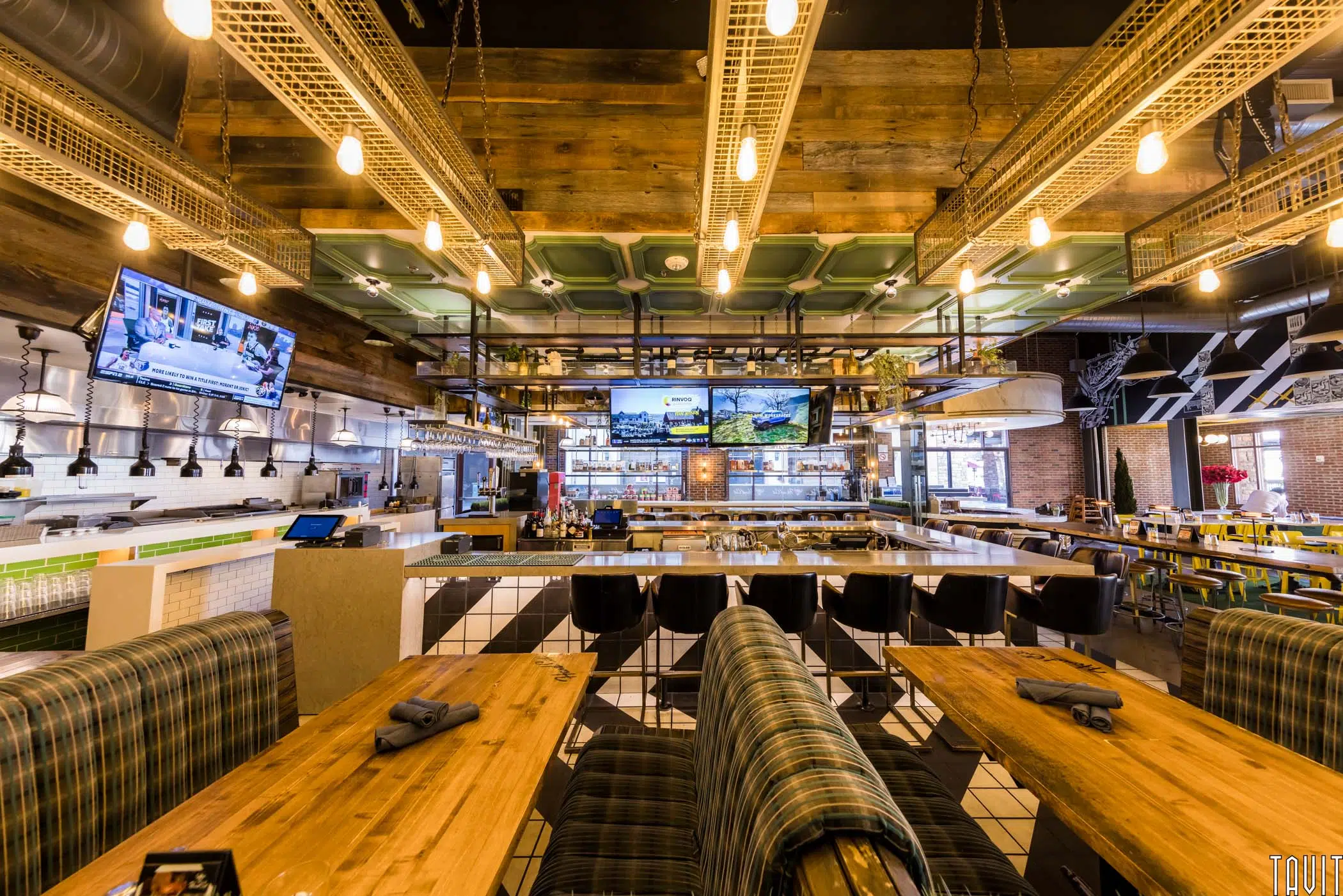A modern restaurant interior with wooden tables, plaid-cushioned seating, a central bar area, and multiple flat-screen TVs—ideal for hosting a corporate event or seminar amid exposed beams, tiled surfaces, and industrial decor.