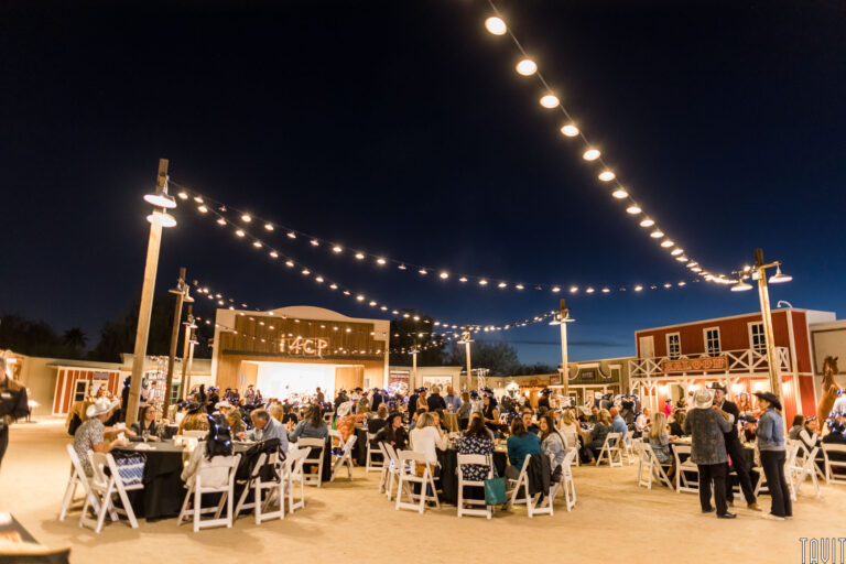 A large outdoor corporate event with people seated at round tables under string lights, surrounded by Western-style buildings and a lit stage in the background. The dark sky enhances the festive seminar atmosphere.