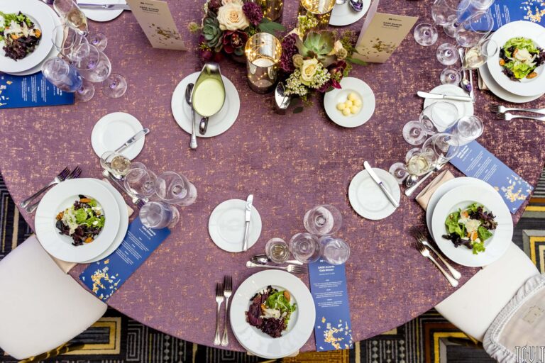 Overhead view of a round table set for a corporate event, featuring salads on white plates, menus, wine glasses, silverware, and elegant floral centerpieces on a textured purple tablecloth.